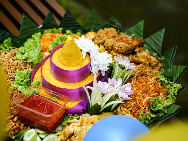Tumpeng nasi kuning dengan hiasan sayur dan aneka lauk di sekelilingnya