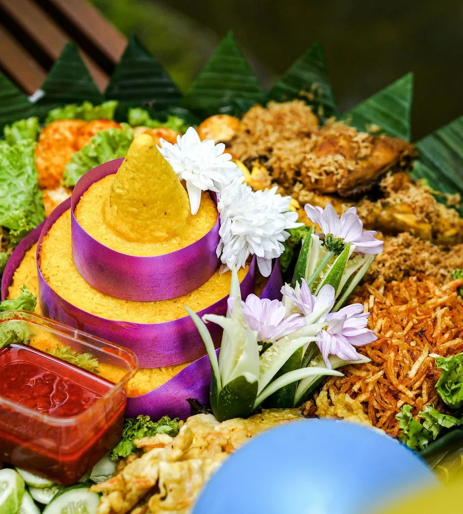 Tumpeng nasi kuning dengan hiasan sayur dan aneka lauk di sekelilingnya